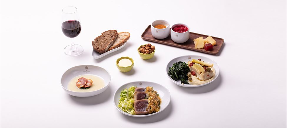 Composition with several elements: a tall glass of red wine, a bowl with two slices of bread, a butter dish, a bowl with cashew nuts, a plate with two slices of chorizo, a board with cheese, marmalade and two glasses of jam, and two plates - one with lettuce, steak and broa (corn bread) with caramelized onions, and the other with cod, sprouts and mash.