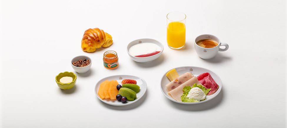 Composition with various elements: a croissant, a butter dish, a bowl of nuts, a jar of peach jam, a bowl of semifreddo, a tall glass of orange juice, two plates - one with grapes, orange, kiwi and sliced strawberries, the other with cheese, mortadella and lettuce - and a cup of coffee.