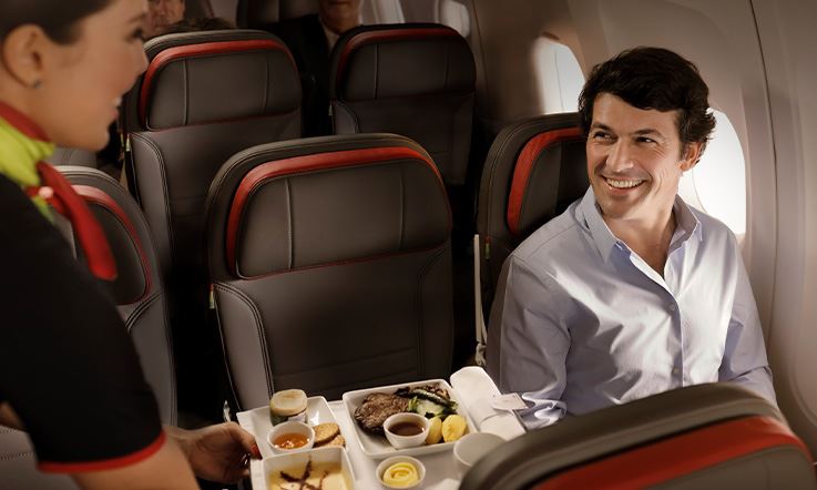 Fotografia de um homem sentado dentro de um avião junto à janela. O homem está a olhar e a sorrir para uma hospedeira que está do lado esquerdo da imagem e que segura numa bandeja com vários pratos de comida.