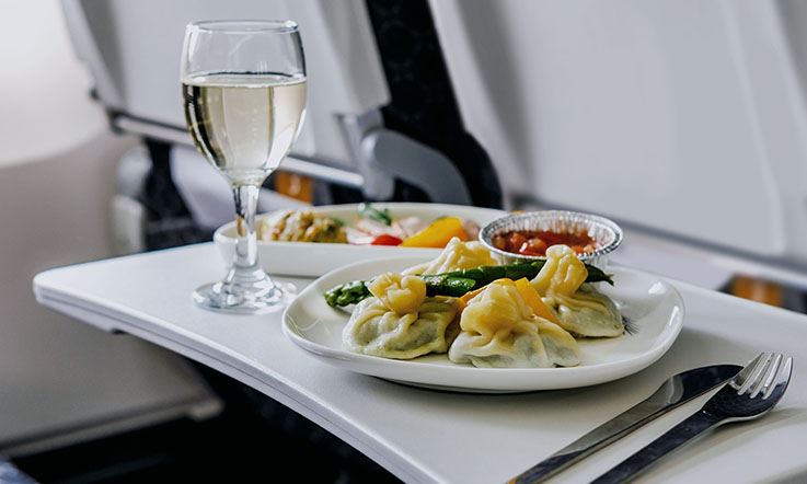 Photography of a meal - two plates of food, a tall glass of white wine and some cutlery - on the side table of an airplane chair.