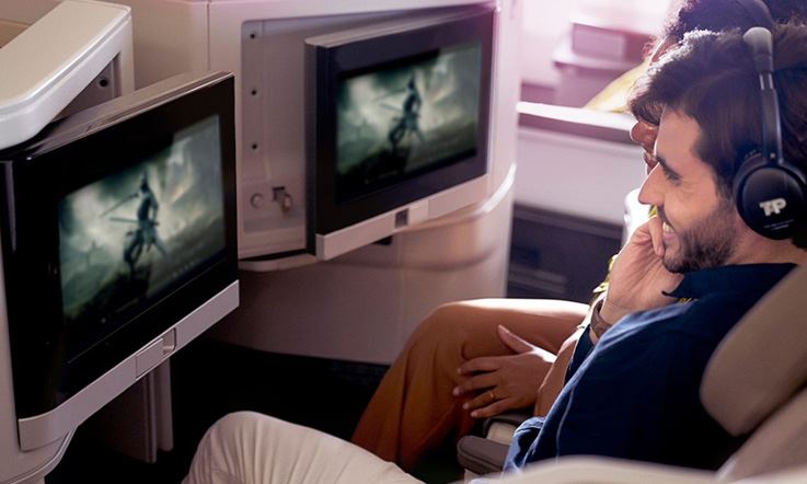 Un couple de passagers souriants, assis en Classe Affaires, dans une ambiance gorgée de lumière naturelle. Ils regardent le même film sur leurs écrans réglables, avec leurs casques audio TAP sur la tête.