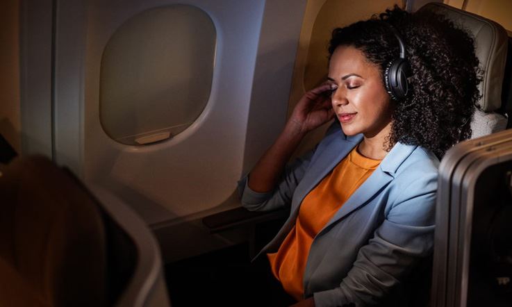 Pasażerka odbywająca lot w wygodnym i przestronnym fotelu klasy biznesowej, w spokojnym i ciemnym otoczeniu, bez kogokolwiek wokół niej. Jest odprężona, ma zamknięte oczy i słucha muzyki w słuchawkach TAP.