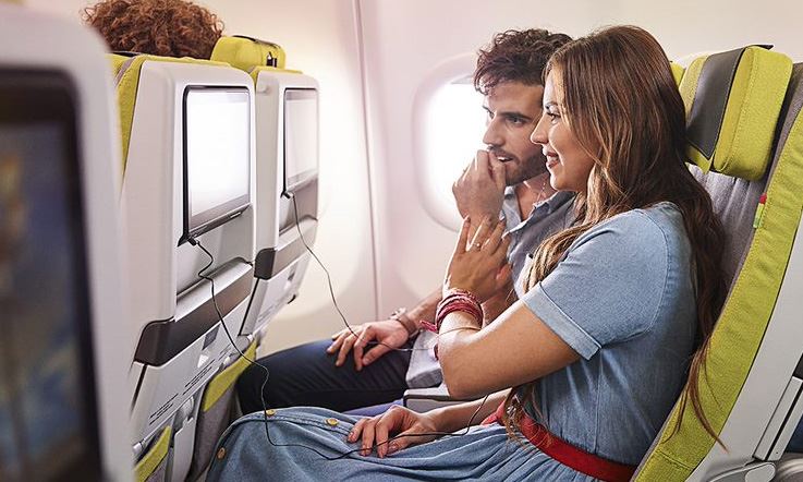 在自然光线充足的环境中，一对乘客坐在绿色的经济舱椅子上。 他们戴着耳机，热切地观看乘客椅背屏幕上的机上电视节目。