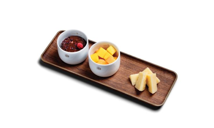 Photography of a wooden tray on which two white bowls rest, flanked on the right by four pieces of cheese. The bowl on the left contains the dessert with chocolate and raspberry textures, and the second bowl contains diced pieces of mango.