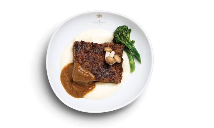 Photography of a white plate with a base of creamy mashed potatoes on which the Maronesa meat terrine rests, topped with three small mushrooms and flanked on the top and right by some Bimi broccoli.