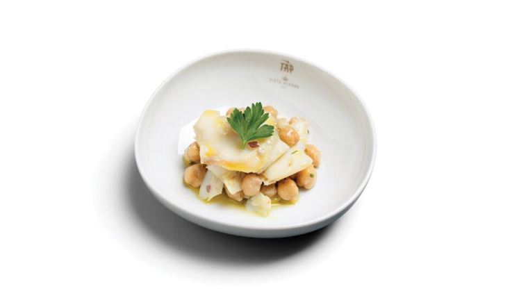 Photography of a white plate with a portion of chickpeas overlaid with some cod chips and a leaf of parsley.