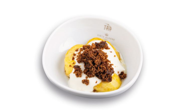 Foto eines weißen Gerichts mit dem TAP-Logo und einem Dessert bestehend aus einer Banane, Frischkäse und einem Kuchen aus Zuckerrohrmelasse.