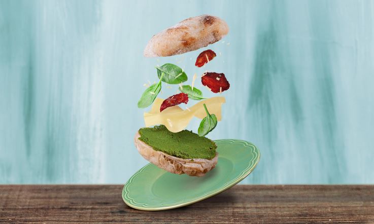 Foto de um prato verde em cima de um tampo de madeira onde se pode ver uma sandes aberta com os ingredientes em suspensão para melhor visualização dos mesmos.
