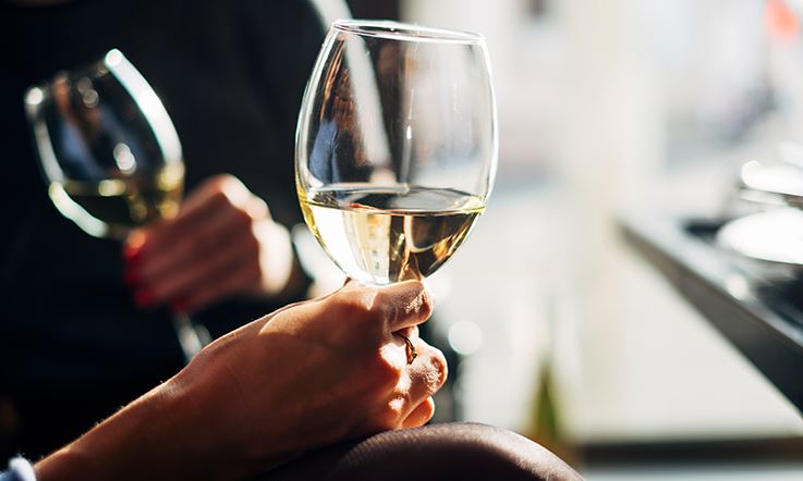 In the foreground, a hand holding a glass of white wine. In the background, more blurred, we see a woman's hands, with nails painted red, holding a glass of white wine, tilting it slightly.