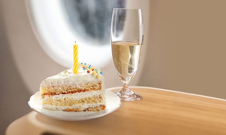 Photo of the inside of an airplane showing, to the right, a slice of cake and a glass of Portuguese sparkling wine standing on a flat surface. In the background there are two airplane windows.