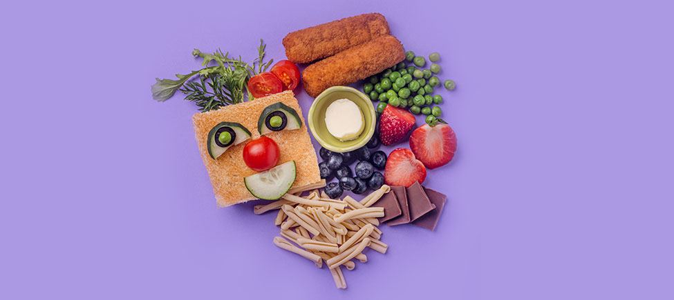 Photo with various ingredients against a purple background: strawberries, peas, blueberries, chocolate, pasta, cherry tomatoes, breaded fish fillets, and a slice of toast with a clown face made of cucumber and tomato.