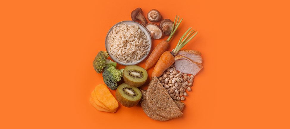 Photo with various ingredients representing a diabetic diet against an orange background: mushrooms, rice, broccoli, carrots, kiwi, cereals, white meat, and other ingredients suitable for this type of diet.