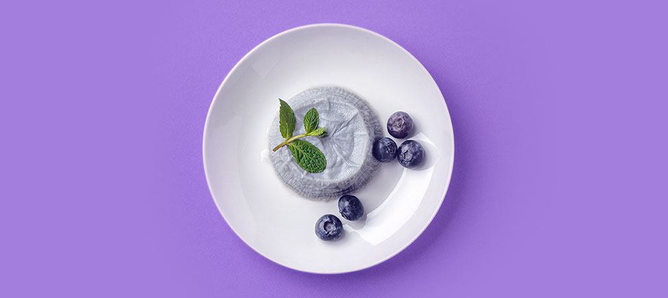 Photo of a white plate with a blueberry mousse topped with a mint leaf and some blueberries on the side, against a purple background.
