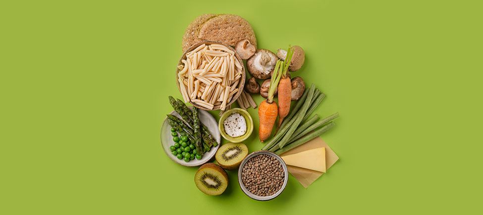 Photo with various ingredients representing a vegetarian diet against a green background: pasta, kiwi, asparagus, peas, carrots, mushrooms, green beans, and other ingredients suitable for this type of diet.