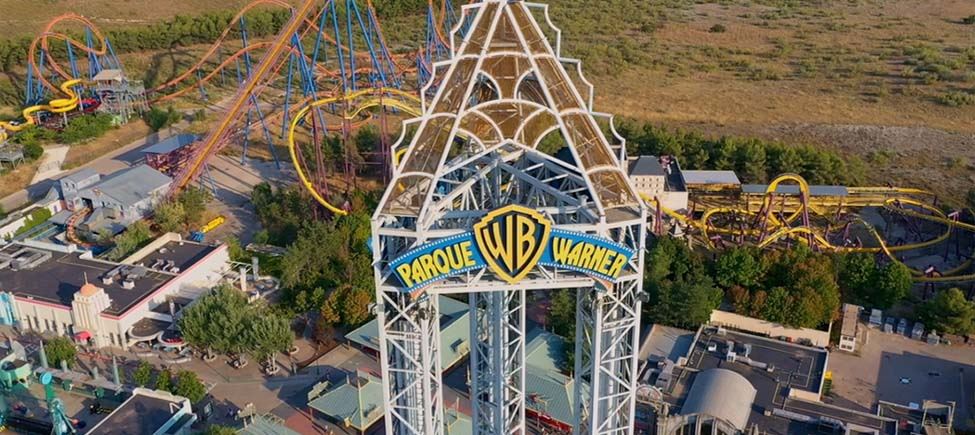 Photo of the top section of an attraction with the Warner Park logo. In the background there are several other attractions from this park and part of an open field with some trees.