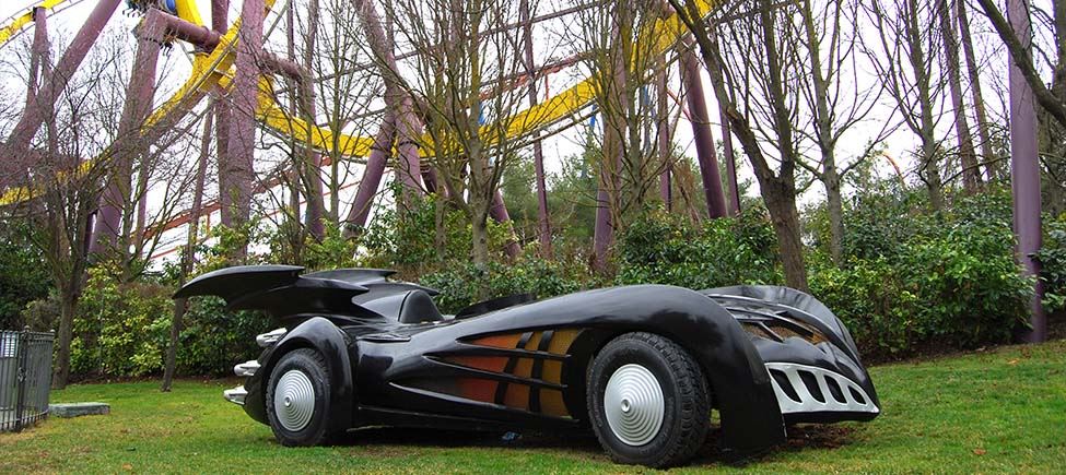 Photo of a car similar to Batman's, in full size. It is black and low slung, with uneven sides and two black wings on the back. It is standing in a garden.
