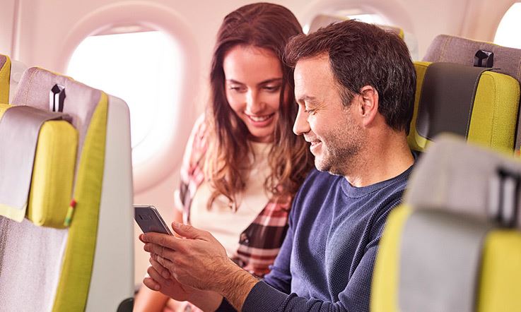 A man and a woman are sitting in the green seats of a TAP plane, while looking at a cell phone and smiling at it.
