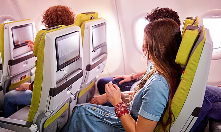 Zdjęcie przedstawia trzech pasażerów siedzących na zielonych fotelach samolotu TAP, patrzących na ekrany znajdujące się na oparciach foteli przed nimi. Pasażer w pierwszym rzędzie dotyka ekranu prawą ręką, podczas gdy pasażerowie w tylnym rzędzie siedzą wygodnie na odchylonych do tyłu fotelach z założonymi słuchawkami i patrzą w ekrany.